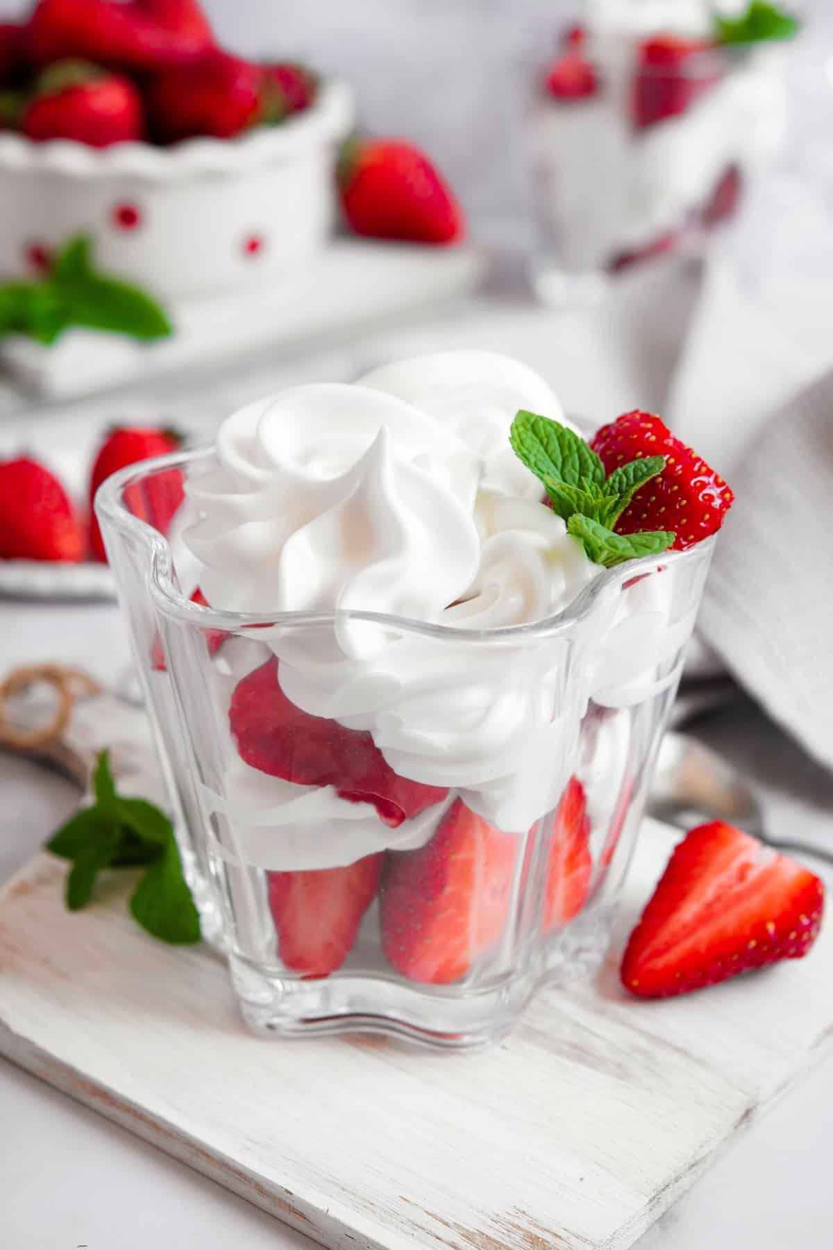 Aquafaba whipped cream piped into a glass cup over fresh strawberries, showing glossy stiff peaks with mint garnish.