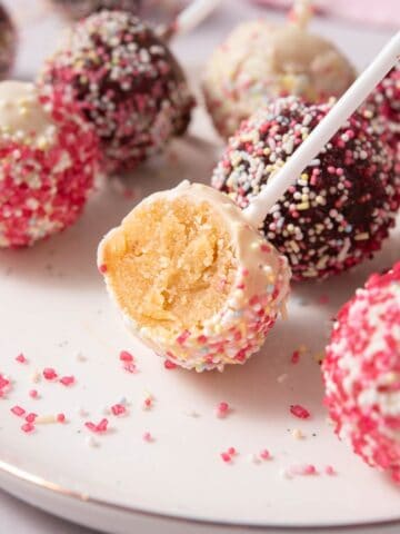 Vegan cake pops coated in vegan white chocolate and vegan dark chocolate with sprinkles on a plate, one bitten open showing vanilla cake