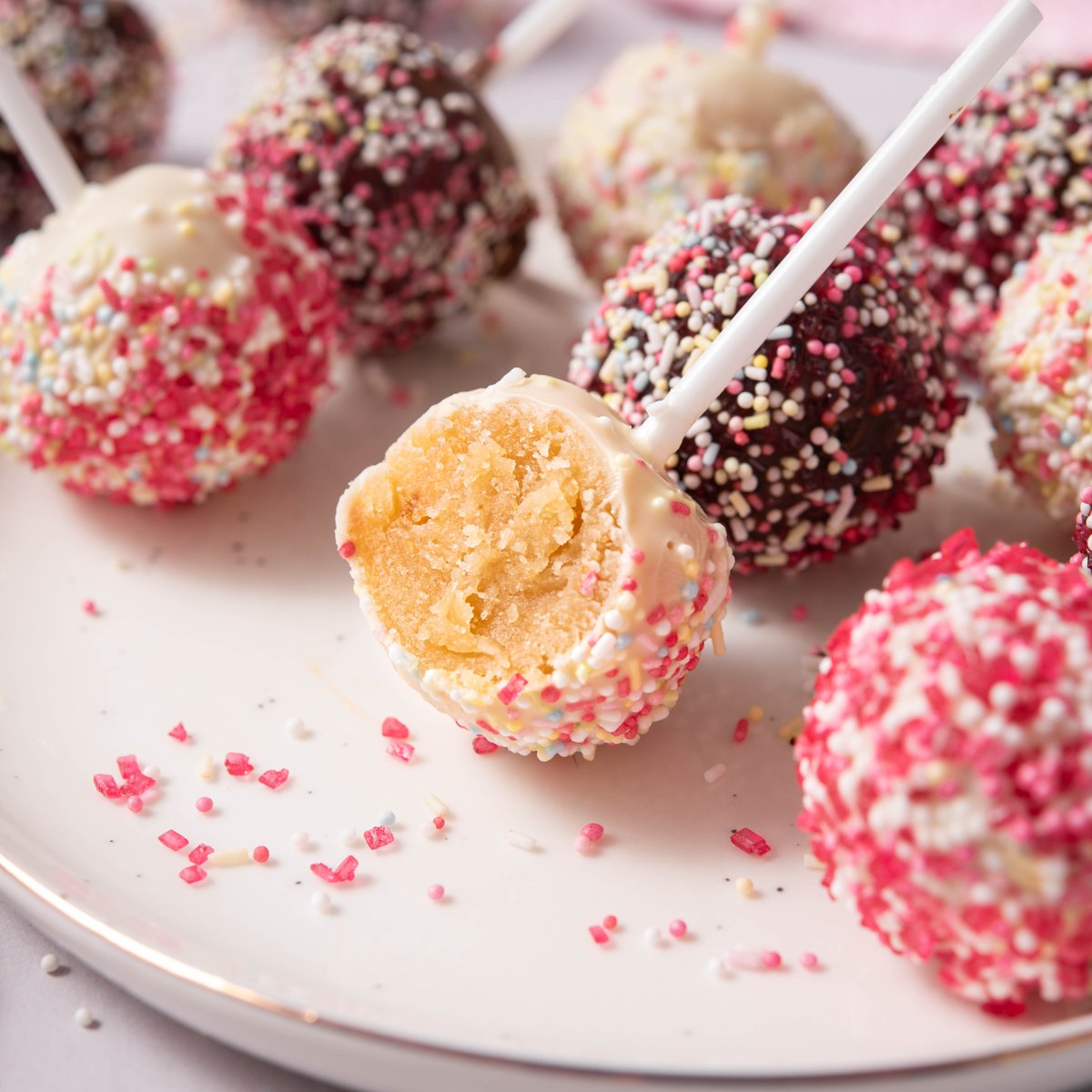 Vegan cake pops coated with vegan white chocolate and vegan dark chocolate, sprinkles placed on the plate and one bites open to reveal vanilla cake