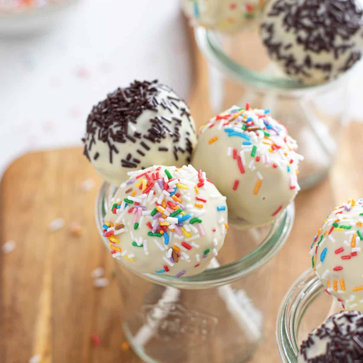 Vegan cake pops dipped in vegan white and dark chocolate, garnished with colorful sprinkles and arranged in small glass jars on a wooden board.