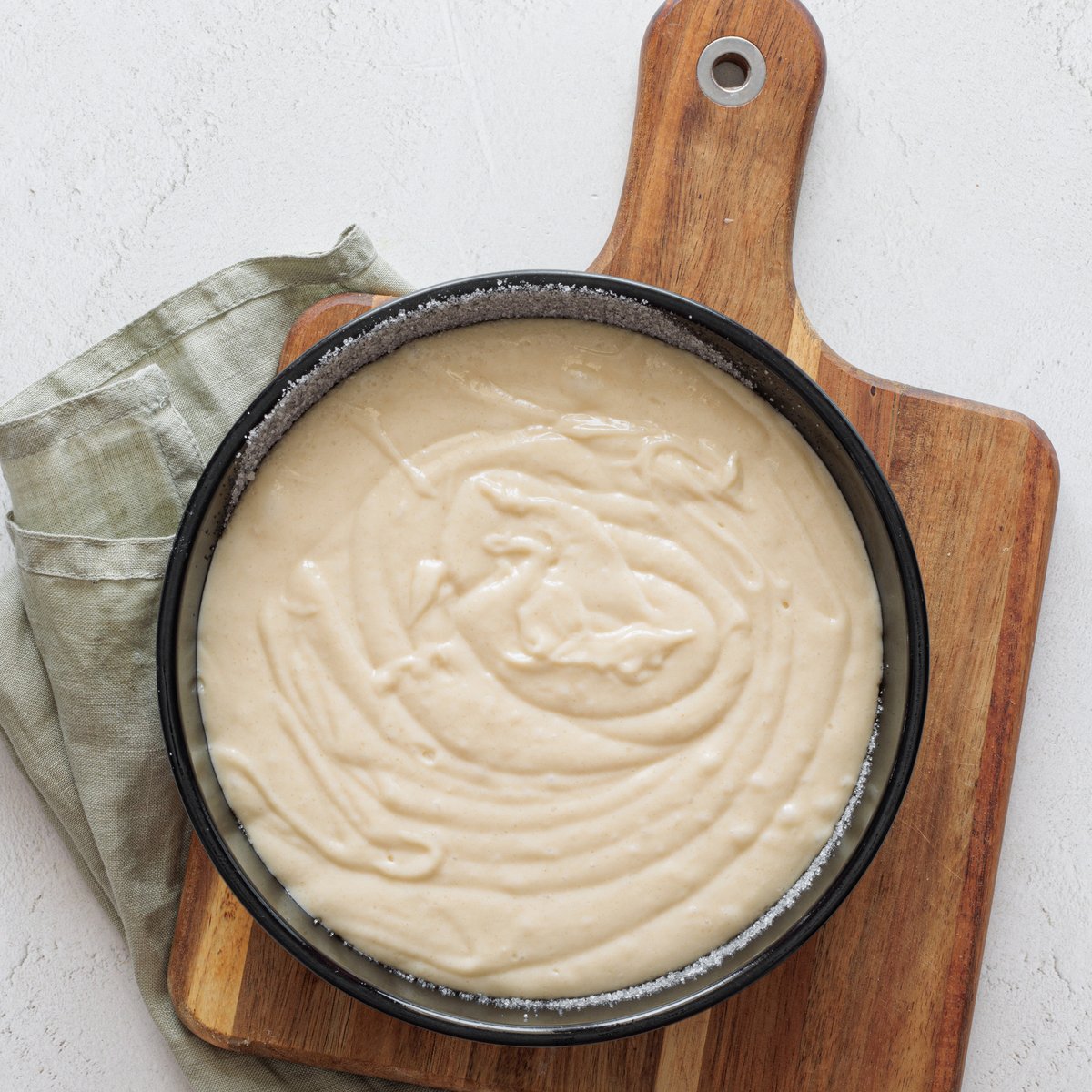On a wooden cutting board with a green cloth napkin, pour the unbaked vegan cake pop batter into a springform pan topped with a bread crust.