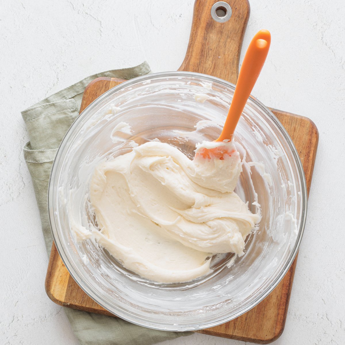 Creamy vegan frosting made with vegan butter and powdered sugar in a glass bowl and an orange spatula on a wooden board.
