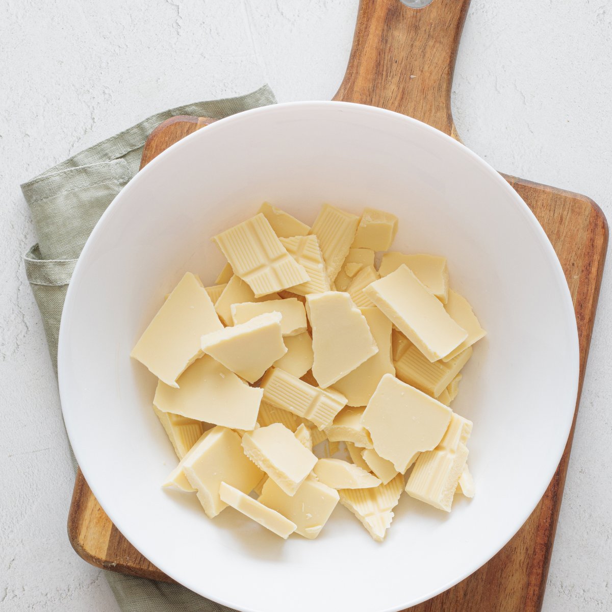 Broken chunks of vegan white chocolate in a white bowl on a wooden cutting board with a green linen napkin