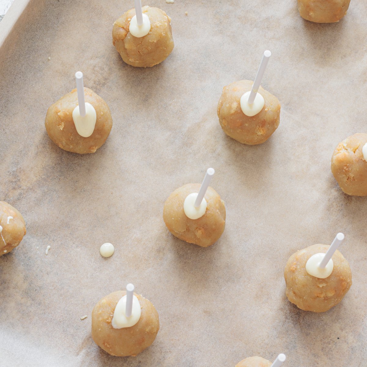 Balls of vegan cake pop batter with cake pop sticks inserted and melted vegan white chocolate on a baking sheet lined with parchment paper.