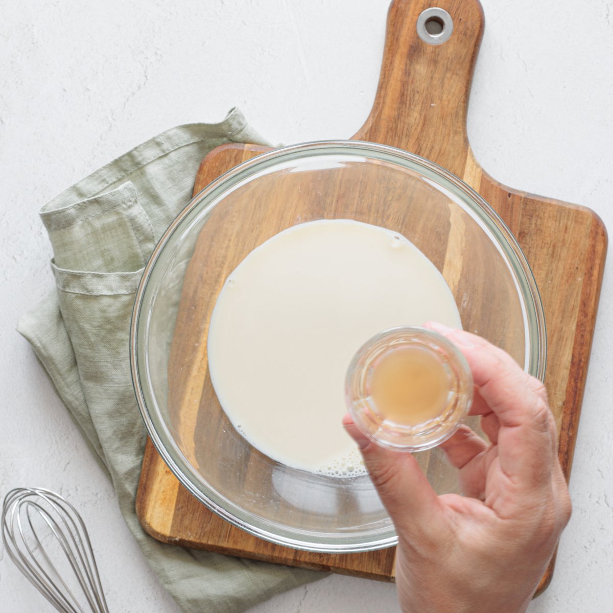 Using a nearby whisk, pour white vinegar by hand into a glass bowl of unsweetened plant-based milk on a wooden board.