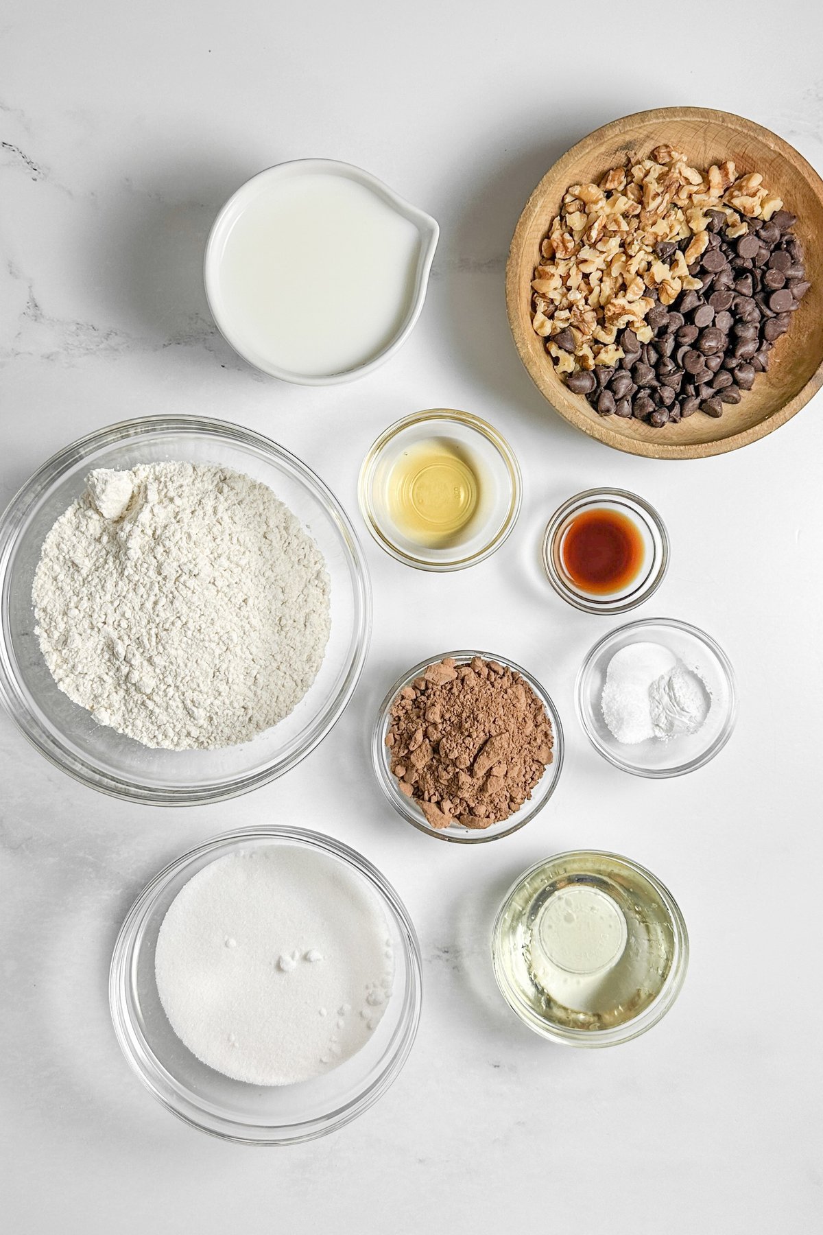 Chocolate loaf cake ingredients in glass bowls on marble: flour, cocoa, sugar, oil, milk, vanilla, walnuts, and chocolate chips