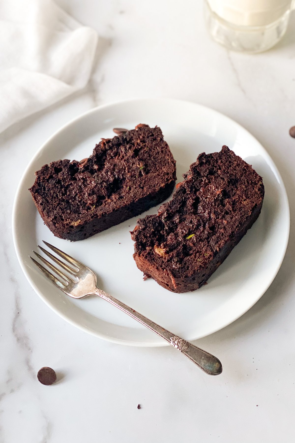 Two slices of chocolate loaf cake on a white plate with a vintage silver fork, showing fudgy crumb and chocolate chips on marble
