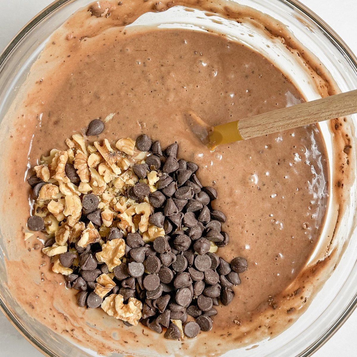 Chocolate loaf cake batter in a glass bowl with chopped walnuts and chocolate chips being folded in with a wooden spoon