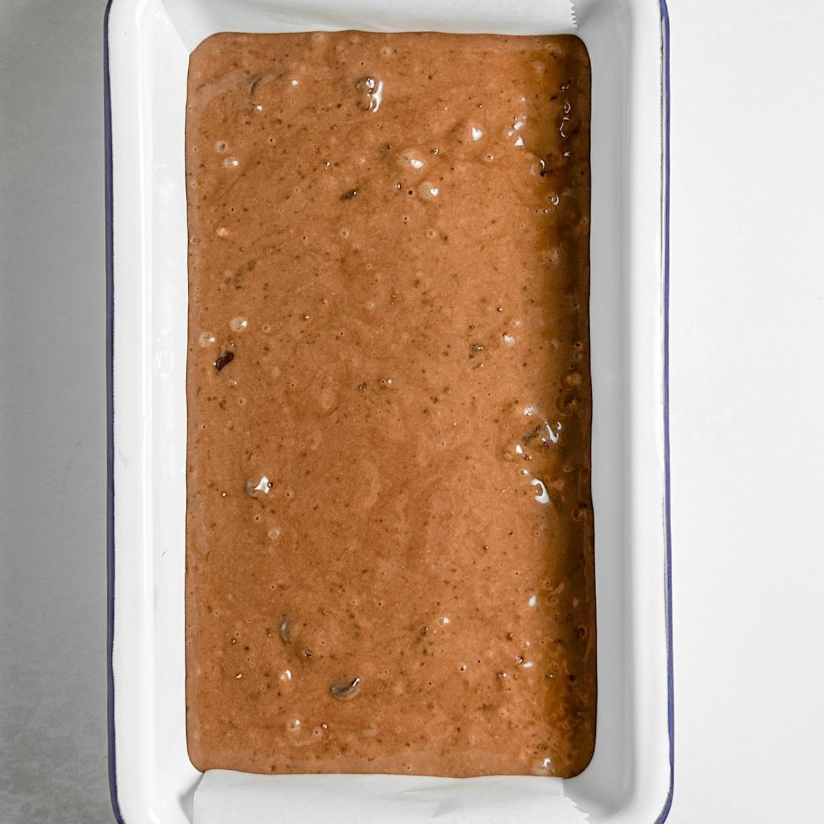 Chocolate loaf cake batter poured into a white enamel loaf pan lined with parchment paper, ready to bake