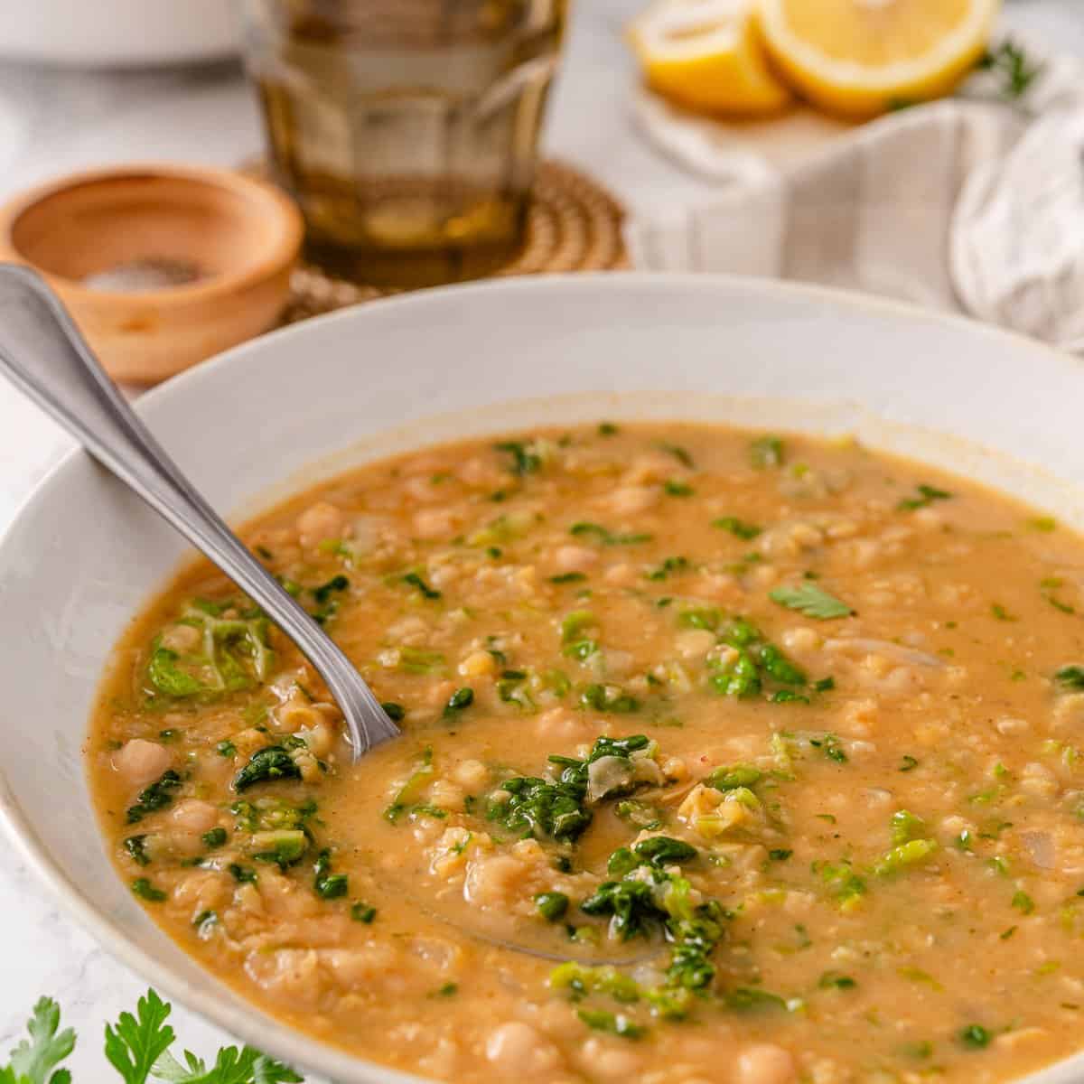 Chickpea lentil soup in a white bowl with red lentils, chickpeas, chopped kale, and a spoon, beside lemon wedges on marble