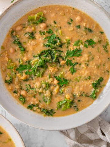 Chickpea lentil soup in a rustic white ceramic bowl with red lentils, chickpeas, chopped kale, and fresh parsley on marble