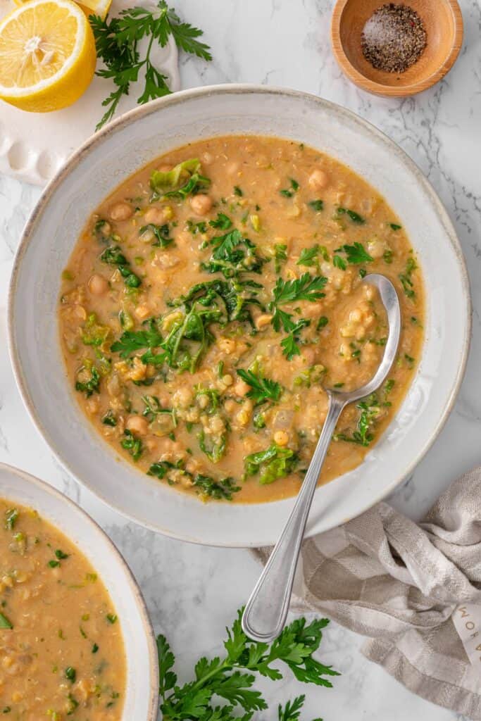Bowl of Chickpea Lentil Soup with red lentils, chickpeas, chopped kale, and fresh parsley garnish, served with a silver spoon on marble.