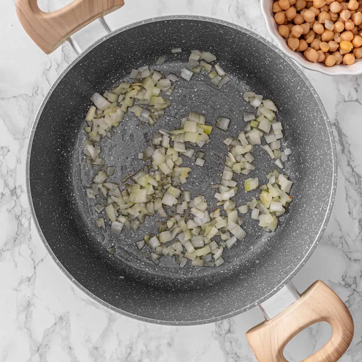 Finely diced onion saut&eacute;ing in a dark nonstick skillet beside a white bowl of drained and rinsed chickpeas for chickpea lentil soup.