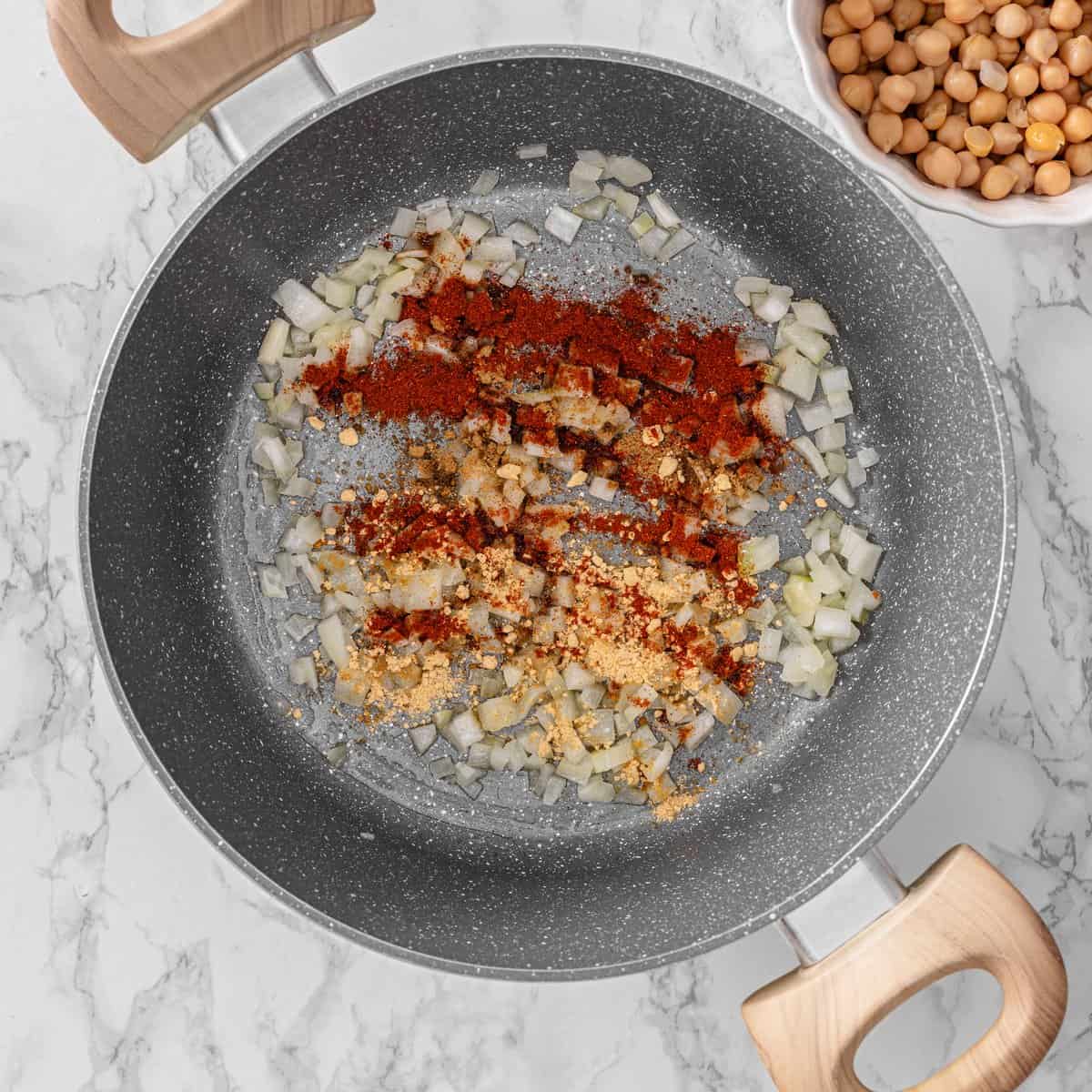 Diced onion cooking in a gray nonstick skillet with smoked paprika and ginger powder added, chickpeas in a bowl nearby.