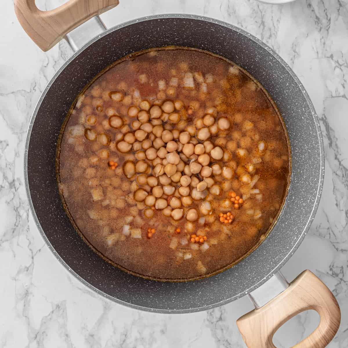 Chickpea lentil soup simmering in a pan with red lentils, chickpeas, diced onion, and spiced vegetable broth on marble