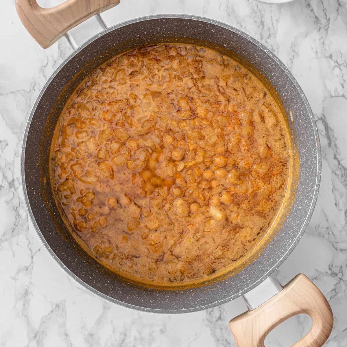 Chickpea lentil soup simmering in a large pan with red lentils, chickpeas, and spiced broth bubbling on a marble surface.