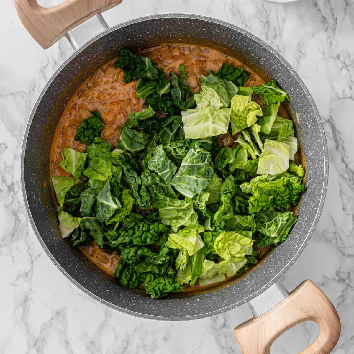 Chopped kale added to simmering red lentil and chickpea soup base in a large gray pan on marble surface.