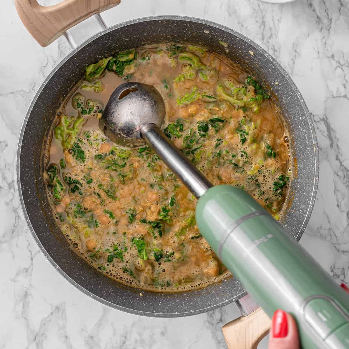 Chickpea lentil soup simmering in a pan with chopped kale, red lentils, and chickpeas being partially blended with a green immersion blender.