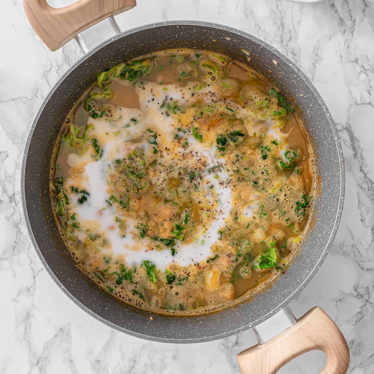 Chickpea lentil soup simmering in a gray pan with coconut cream swirled in, chopped kale, and black pepper on marble surface.
