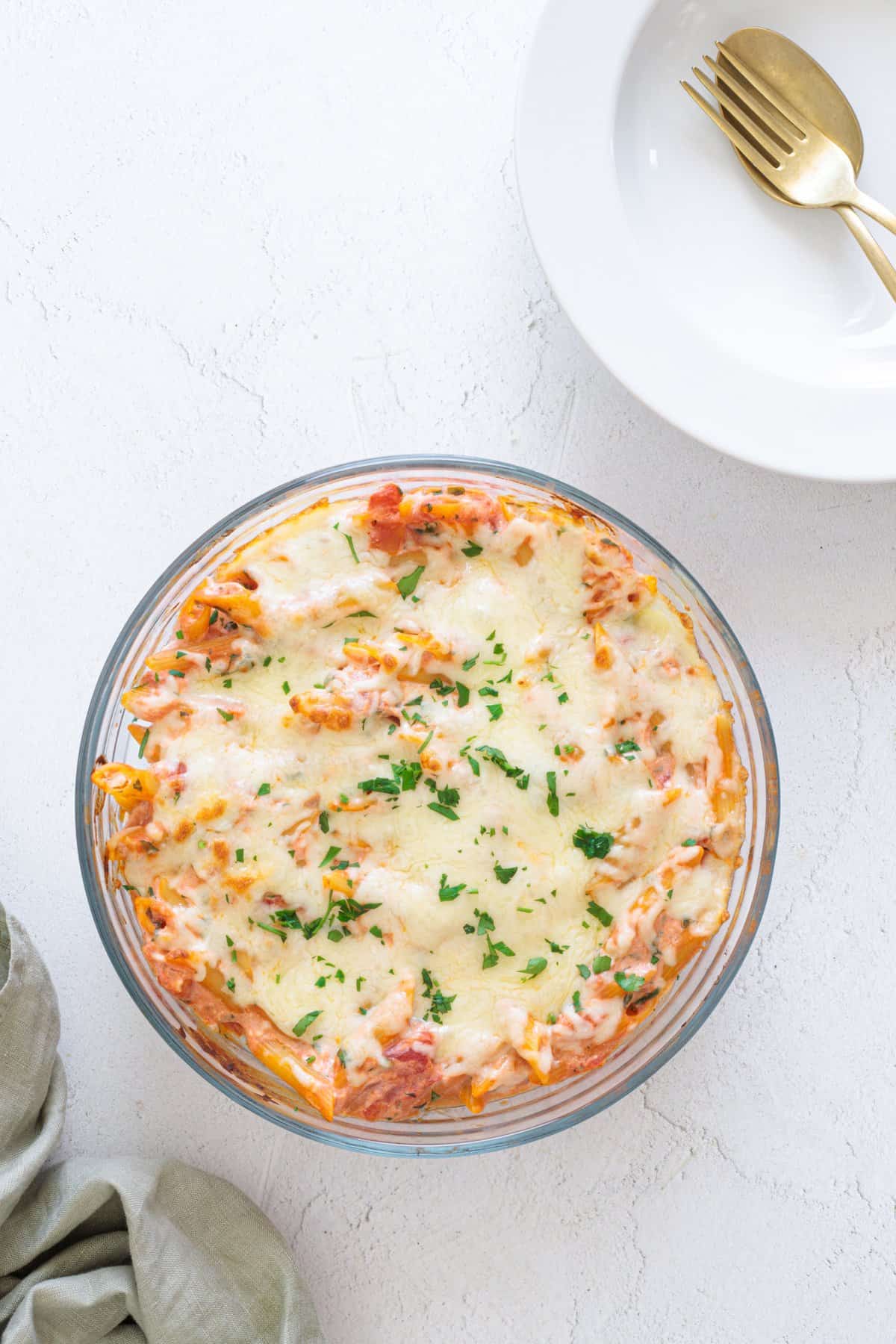 Easy Cheesy Red Lentil Baked Ziti in a round glass baking dish, topped with melted mozzarella and fresh chopped parsley, on a white surface.