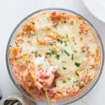 Easy Cheesy Red Lentil Baked Ziti in a round glass baking dish, topped with melted mozzarella and fresh parsley, spoon lifting a cheesy serving