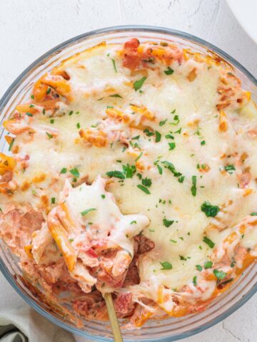 Easy Cheesy Red Lentil Baked Ziti in a round glass baking dish, topped with melted mozzarella and fresh parsley, spoon lifting a cheesy serving