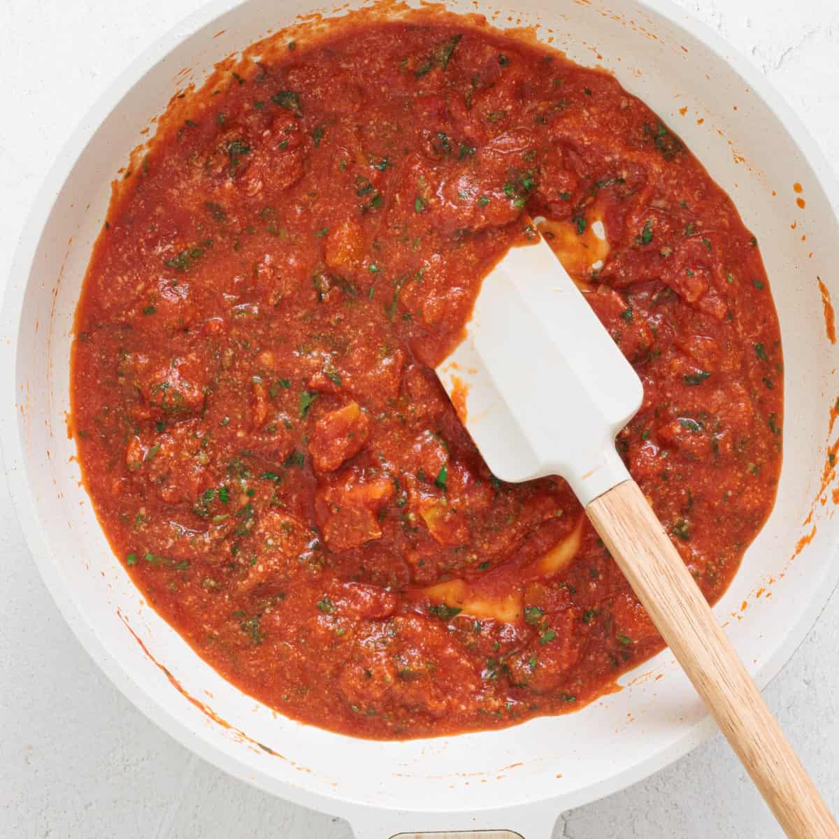 Chunky tomato sauce with fresh herbs simmering in a white skillet, stirred with a white silicone spatula with wooden handle.