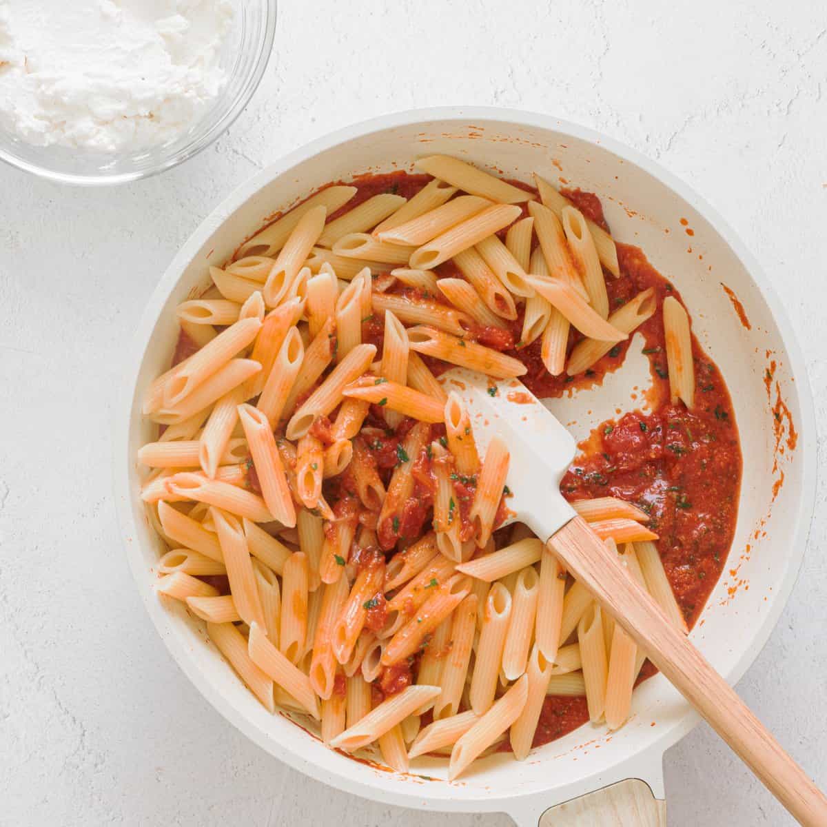 Penne pasta being stirred into chunky red tomato sauce in a white skillet with a wooden spatula, with ricotta in a glass bowl nearby.