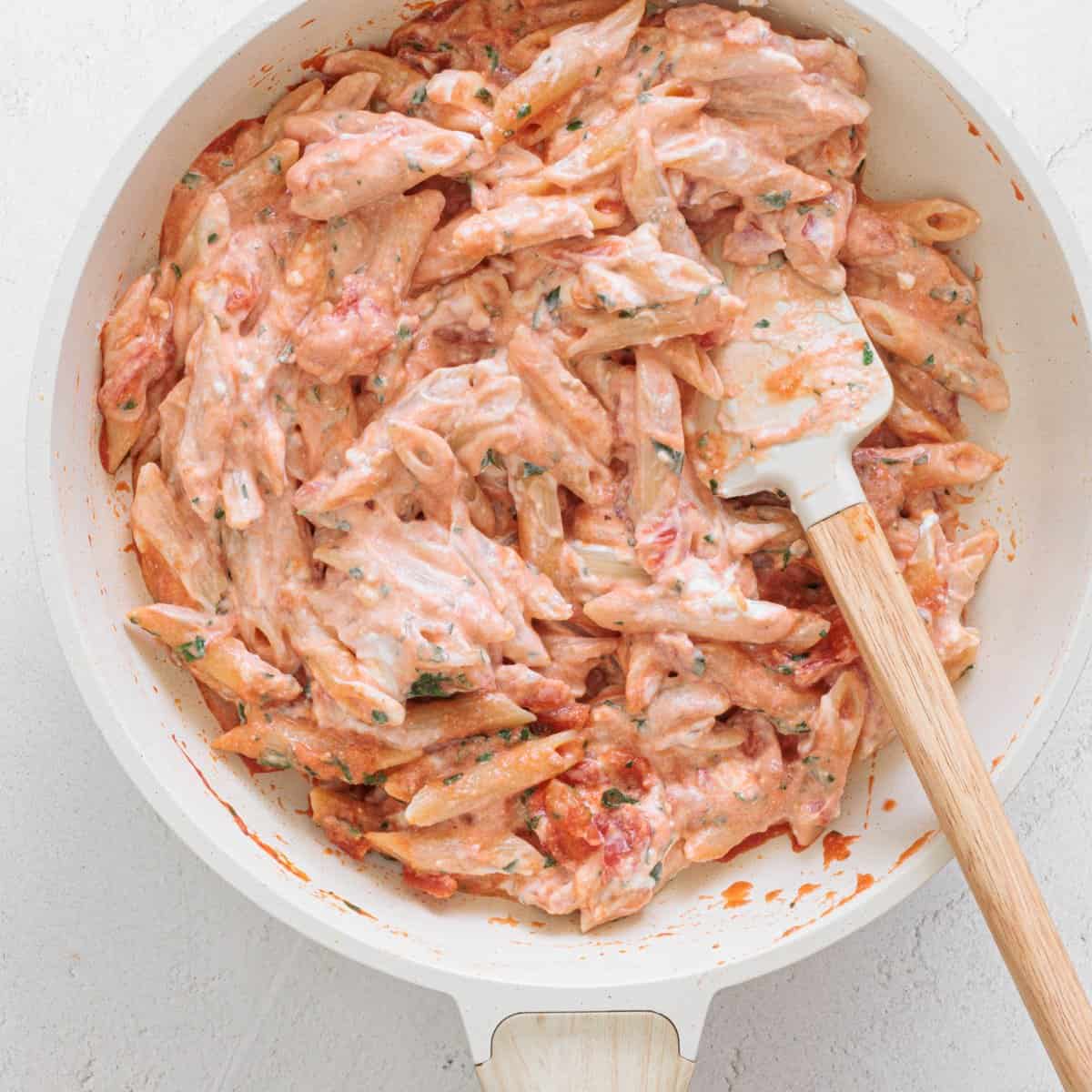 Creamy tomato sauce coating penne pasta being stirred in a white skillet for Easy Cheesy Red Lentil Baked Ziti
