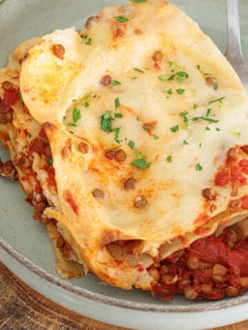 Serving of vegan lentil lasagna on a grey ceramic plate showing layers of pasta, lentils in tomato sauce, b&eacute;chamel, and fresh parsley