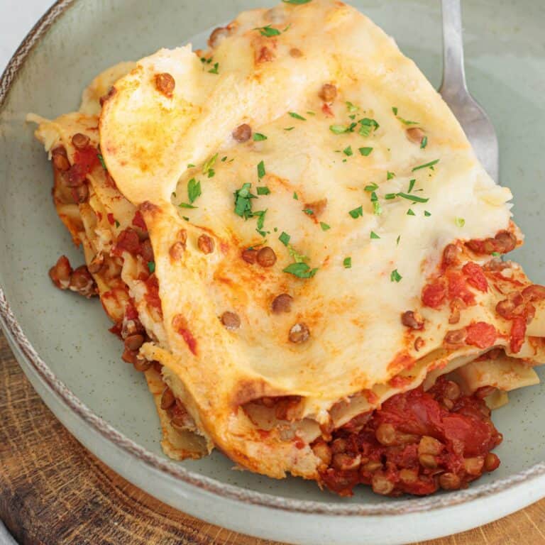 Serving of vegan lentil lasagna on a grey ceramic plate showing layers of pasta, lentils in tomato sauce, béchamel, and fresh parsley