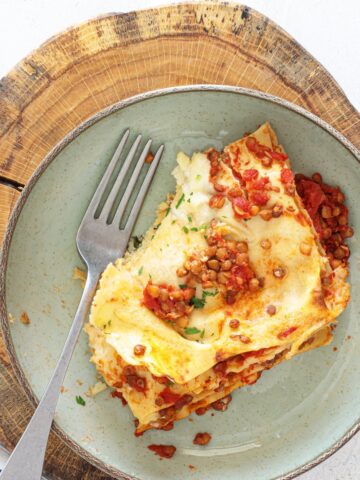 Plated vegan lentil lasagna slice with lentil tomato sauce, b&eacute;chamel, and fresh herbs on a sage green plate atop a wood board with a fork.