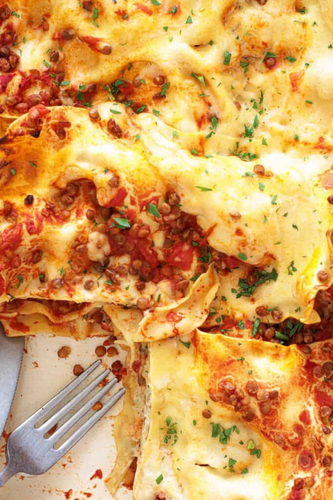 Vegan lentil lasagna in a baking pan with a slice removed, showing lentil tomato sauce layers, béchamel topping, and fresh parsley