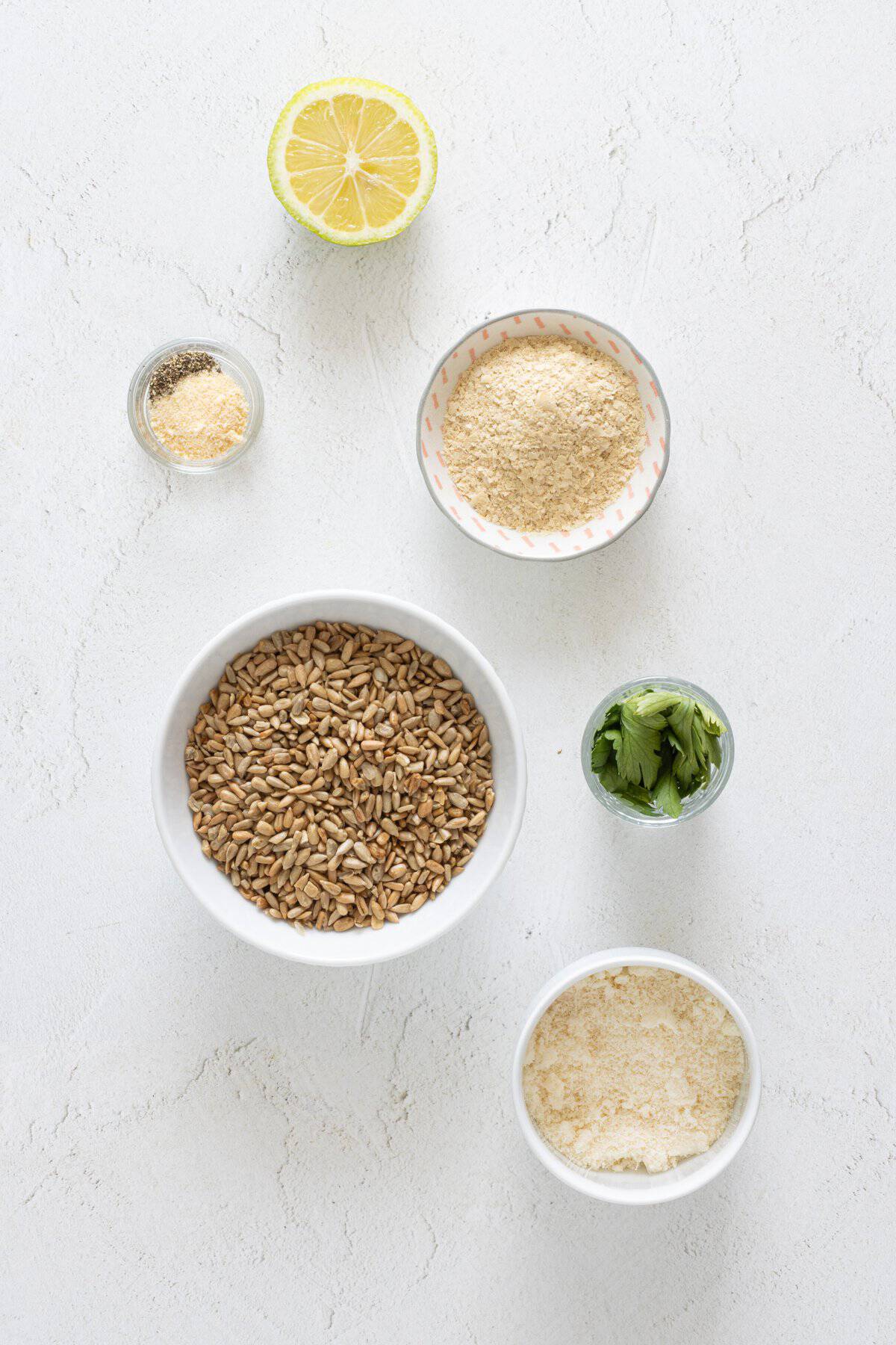 Ingredients for vegan lentil lasagna: sunflower seeds, nutritional yeast, parmesan, fresh parsley, lemon half, and garlic powder on white surface