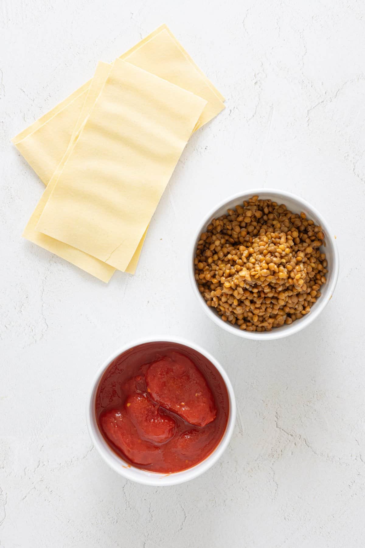 Vegan lentil lasagna ingredients on white surface: lasagna sheets, bowl of lentils, and bowl of crushed tomatoes