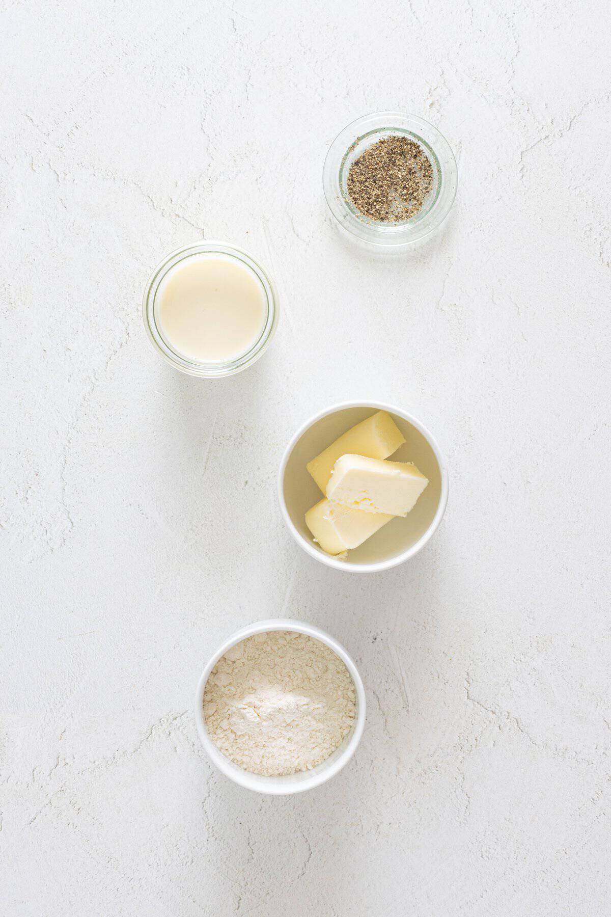 Four bowls on a white surface showing vegan lasagna white sauce ingredients: flour, vegan butter, plant milk, and black pepper