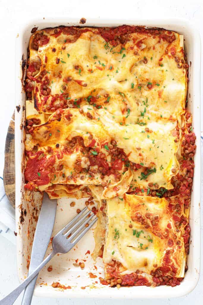 Vegan lentil lasagna in a white baking dish, partially served, with lentil tomato sauce, golden pasta layers, and fresh parsley garnish