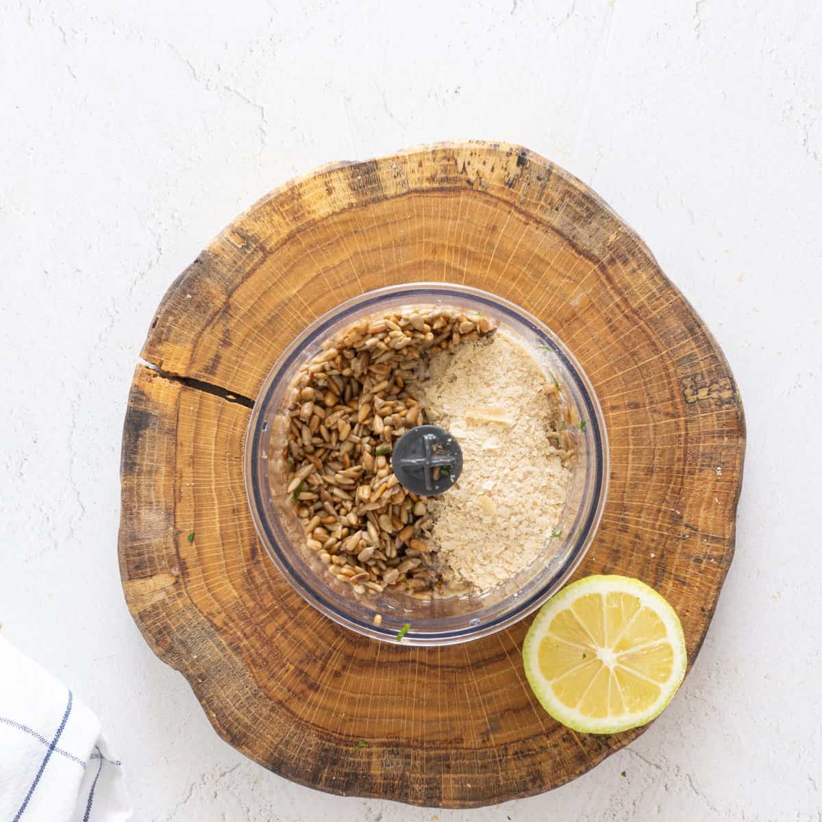 Sunflower seeds and nutritional yeast in a food processor bowl on a wood slice board with a halved lemon beside it