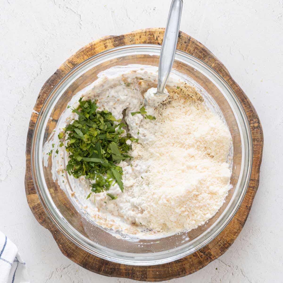 Glass bowl with creamy white bean mixture, chopped fresh parsley, and grated parmesan-style cheese for vegan lentil lasagna on a wood board.
