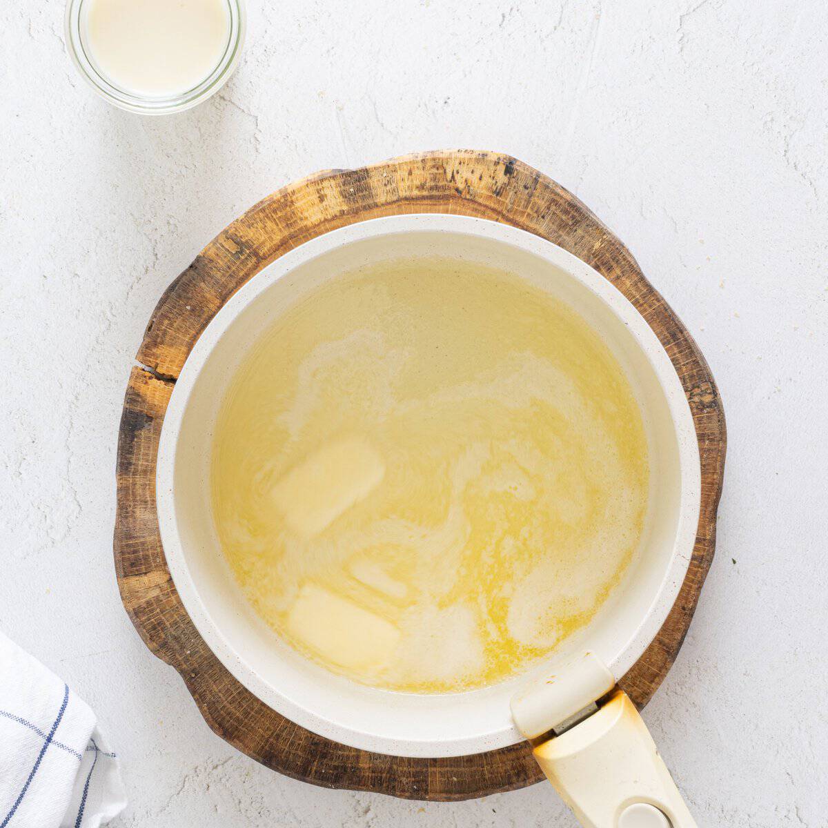 Melting butter in a white saucepan on a wooden trivet beside a small glass of cream for vegan lentil lasagna