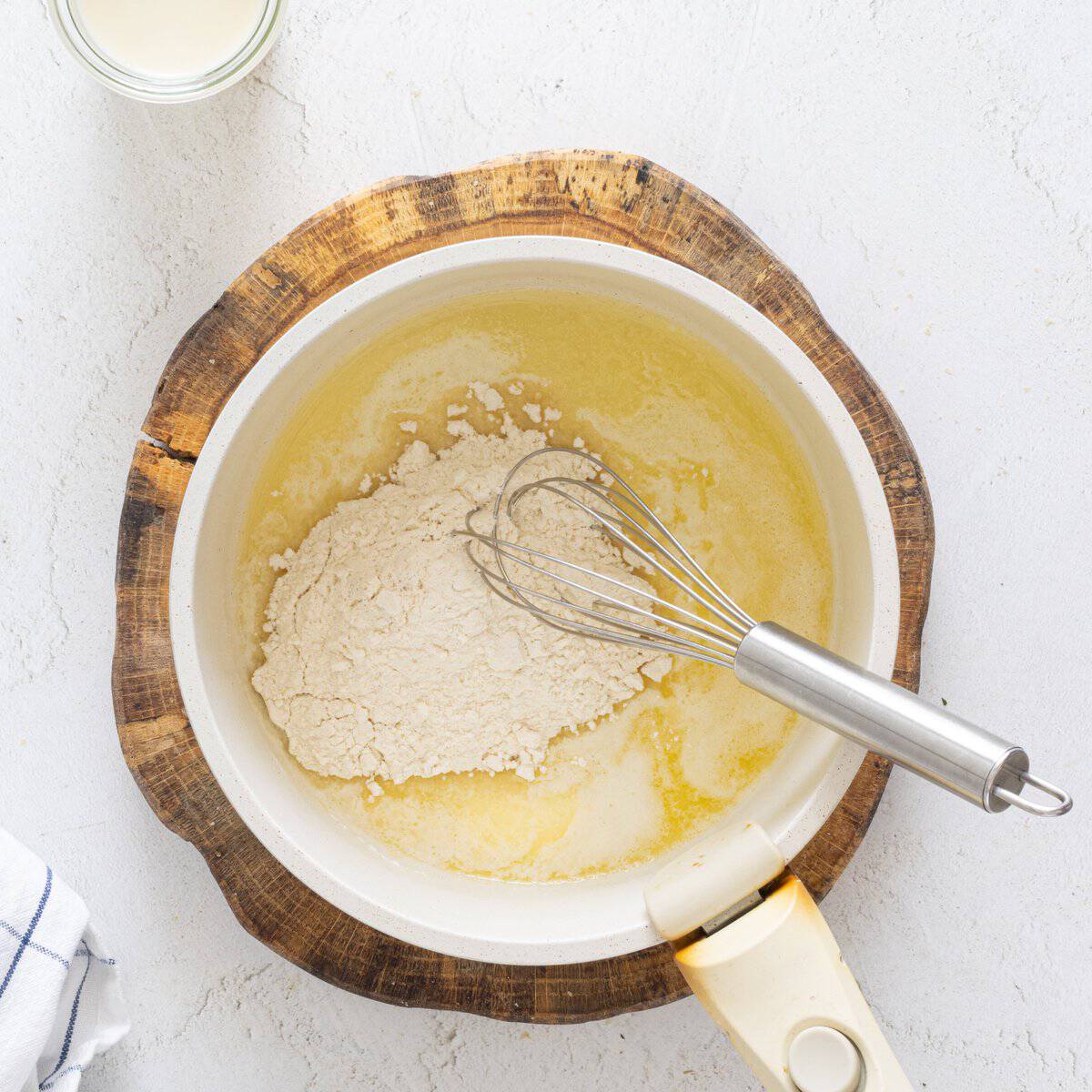 Flour added to melted butter in a white saucepan on a wood board with a whisk, making vegan lentil lasagna béchamel sauce