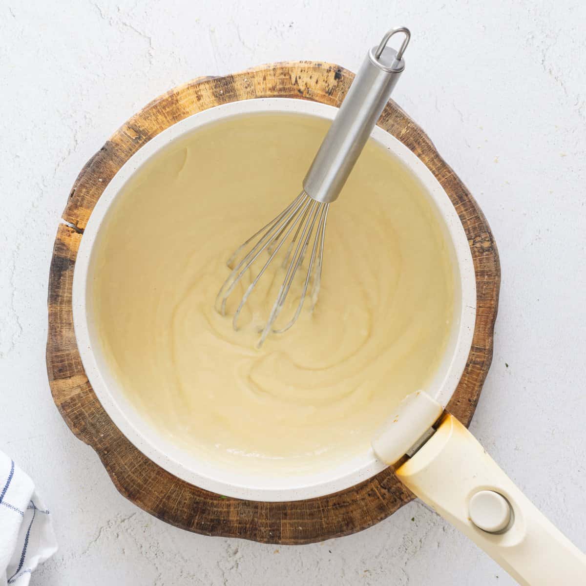 Creamy white sauce being whisked in a saucepan on a wooden board, a step in vegan lentil lasagna preparation.