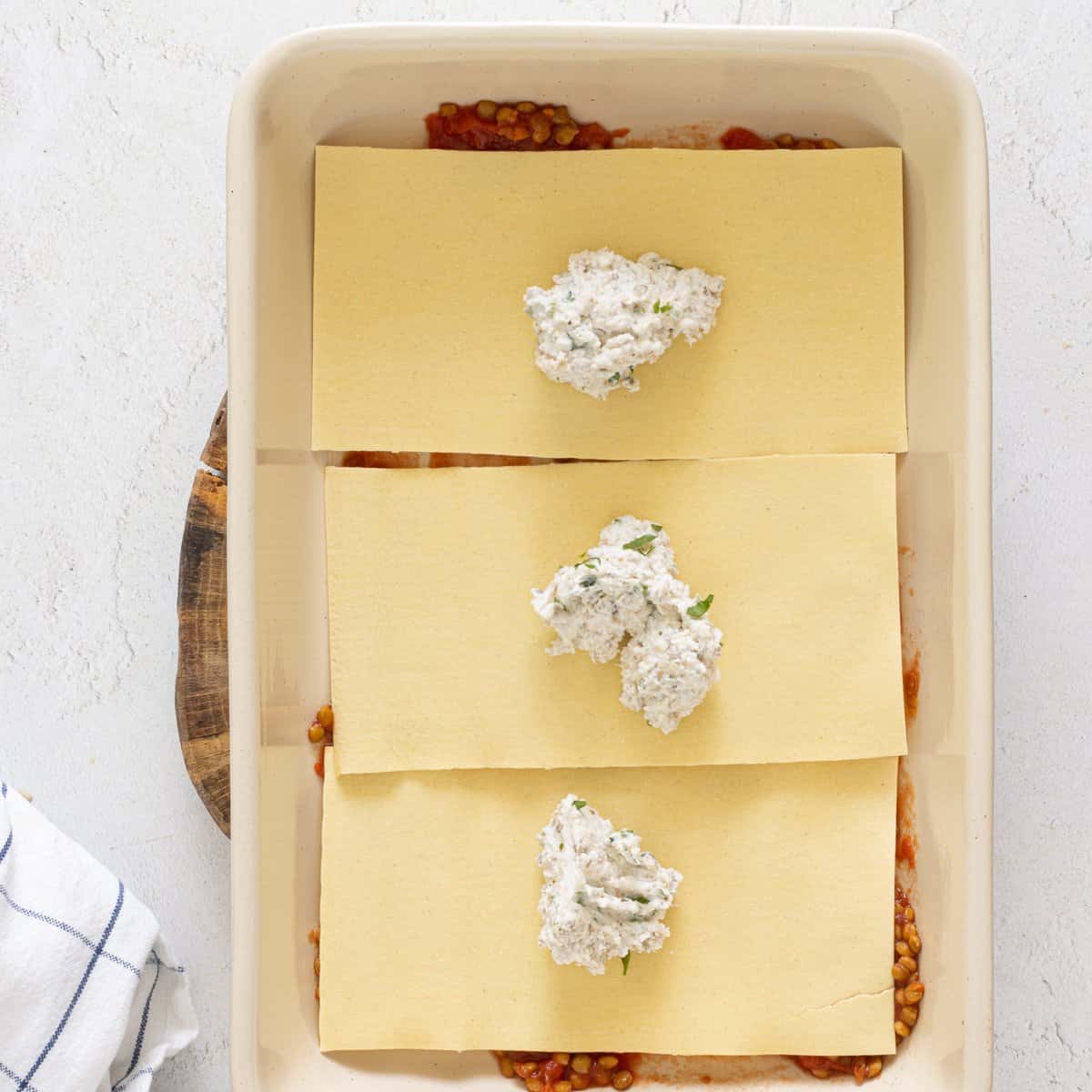 Lasagna noodles layered over lentil tomato sauce in a white baking dish with dollops of vegan ricotta on each noodle