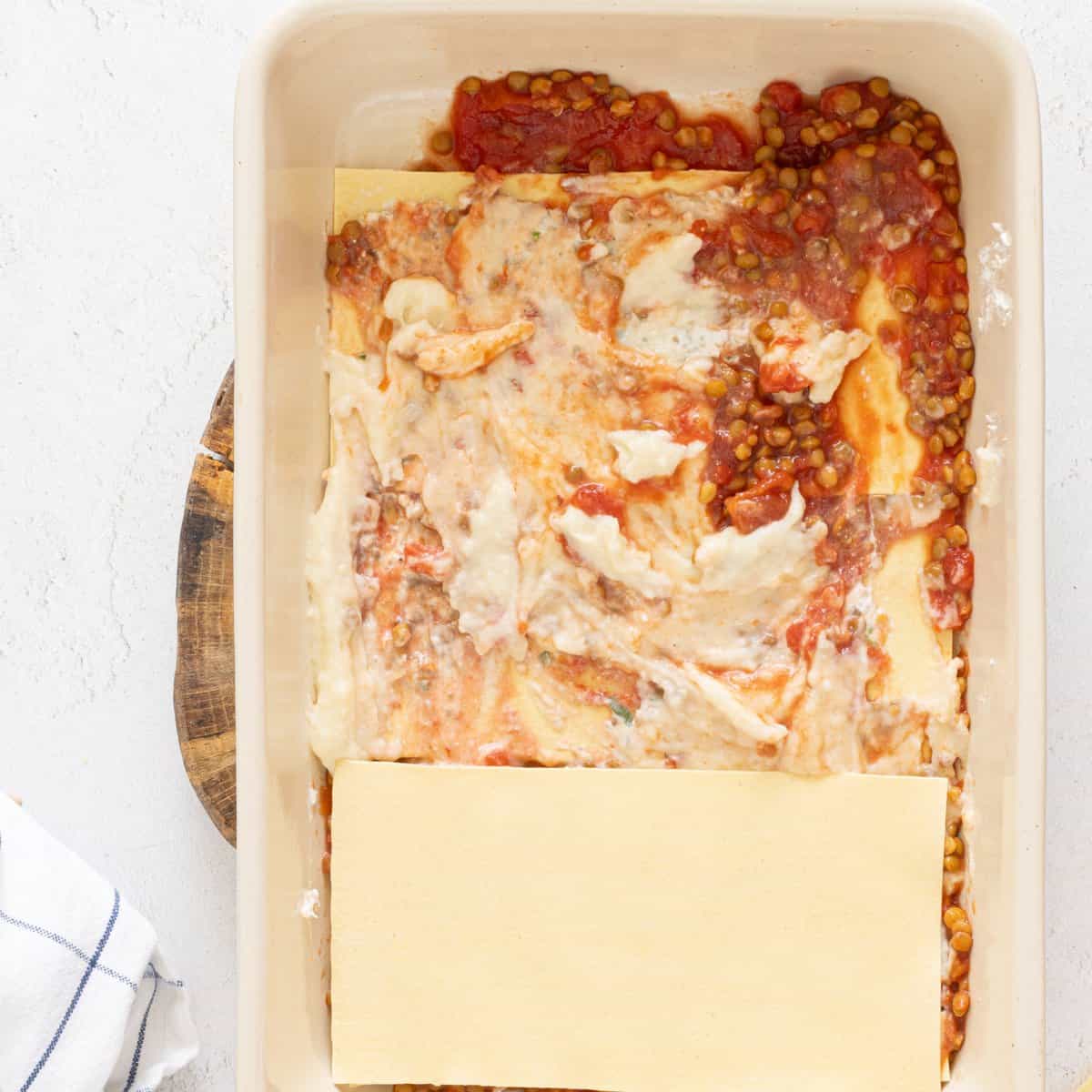 Unbaked vegan lentil lasagna being assembled in a white baking dish with lentil tomato sauce, creamy white layer, and dry lasagna noodles on top.