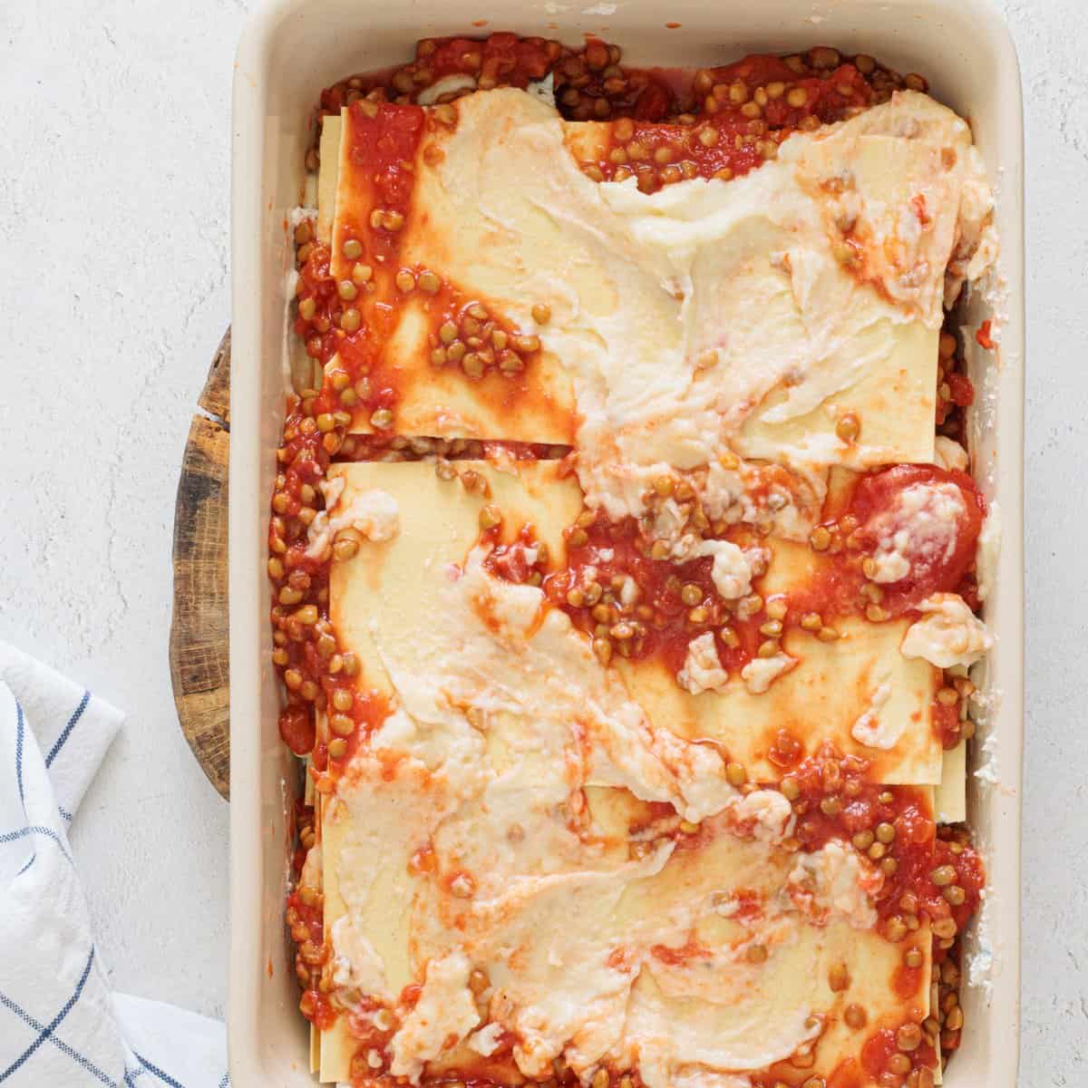 Unbaked vegan lentil lasagna in a white baking dish layered with lasagna noodles, lentil tomato sauce, and dollops of creamy white filling.