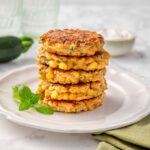 Stack of six golden Zucchini and Corn Fritters packed with corn kernels and grated zucchini on a white plate with fresh herbs