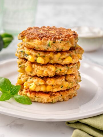 Stack of six golden Zucchini and Corn Fritters packed with corn kernels and grated zucchini on a white plate with fresh herbs