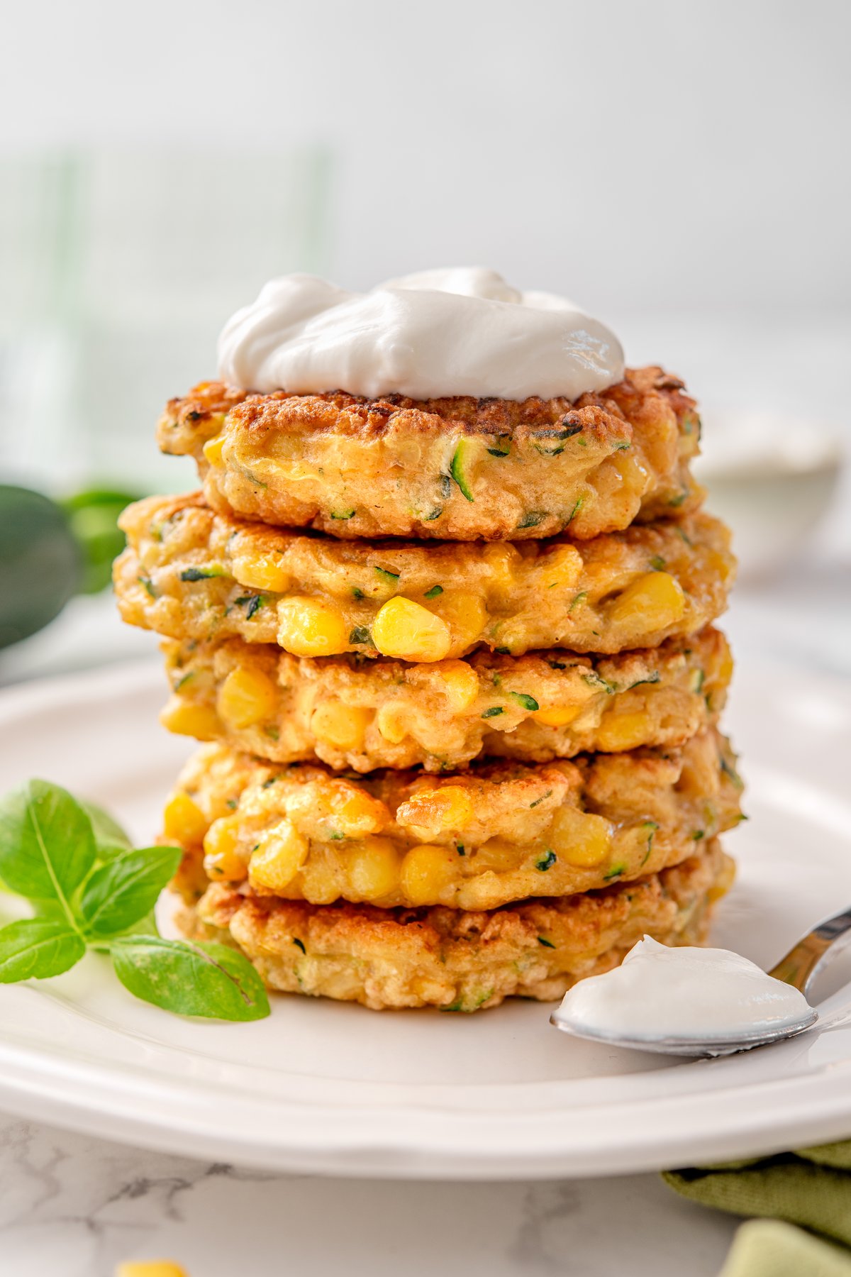 Stack of zucchini and corn fritters with visible corn kernels and grated zucchini, topped with sour cream, on a white plate with fresh basil