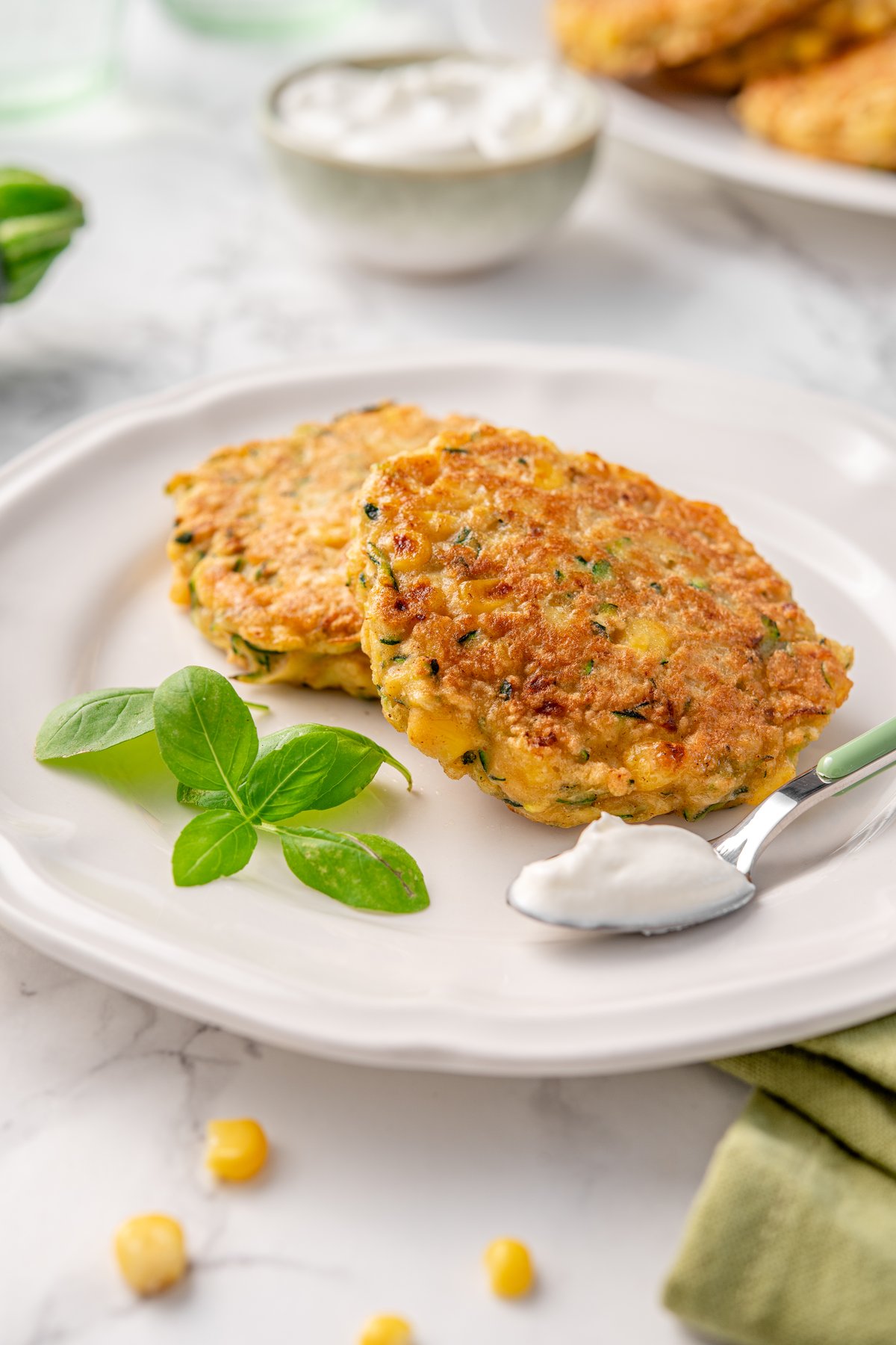 Two golden zucchini and corn fritters on a white plate with fresh basil leaves and a spoon of sour cream on marble