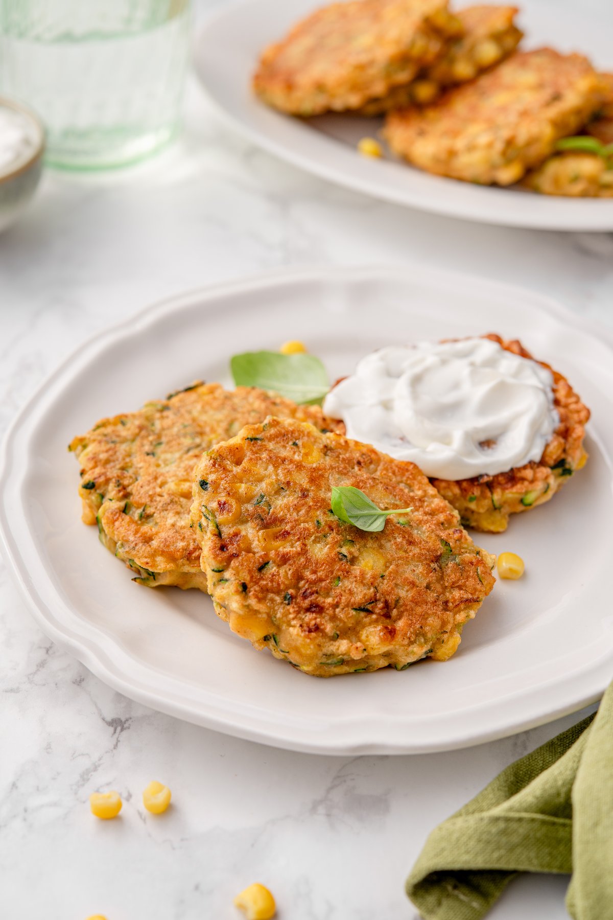 Three golden zucchini and corn fritters on a white plate topped with sour cream and fresh basil leaves on marble